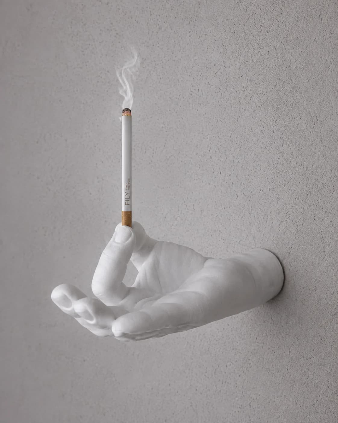 Mano de yeso sosteniendo un incienso FILY encendido contra una pared de revoque gris